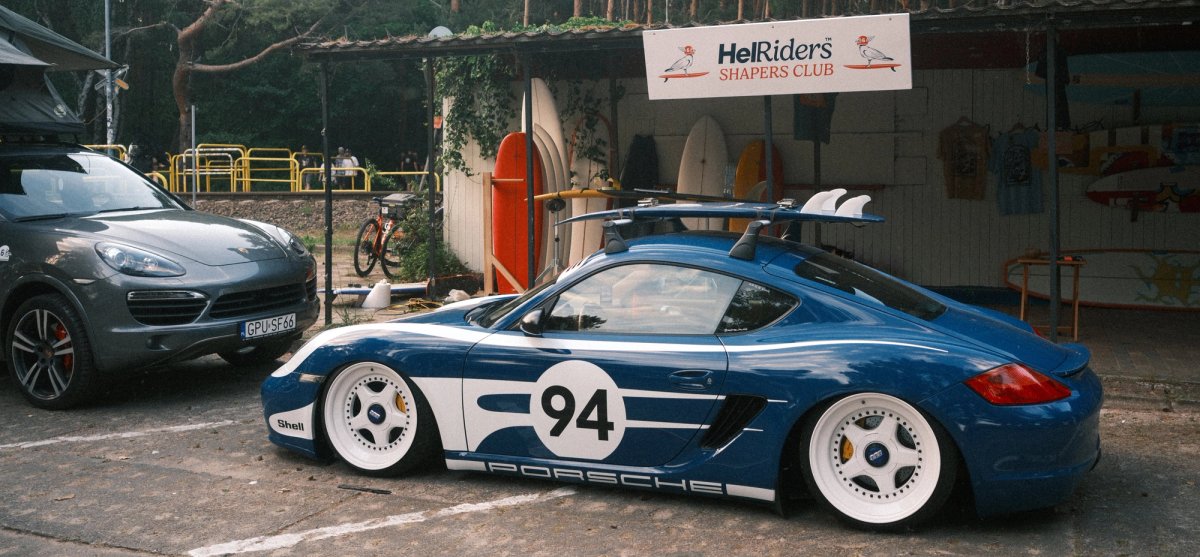 Blue widebody Porsche Cayman number 94 on white BBS wheels in front of the HelRiders Shaper's Club shelter hung with surfboards