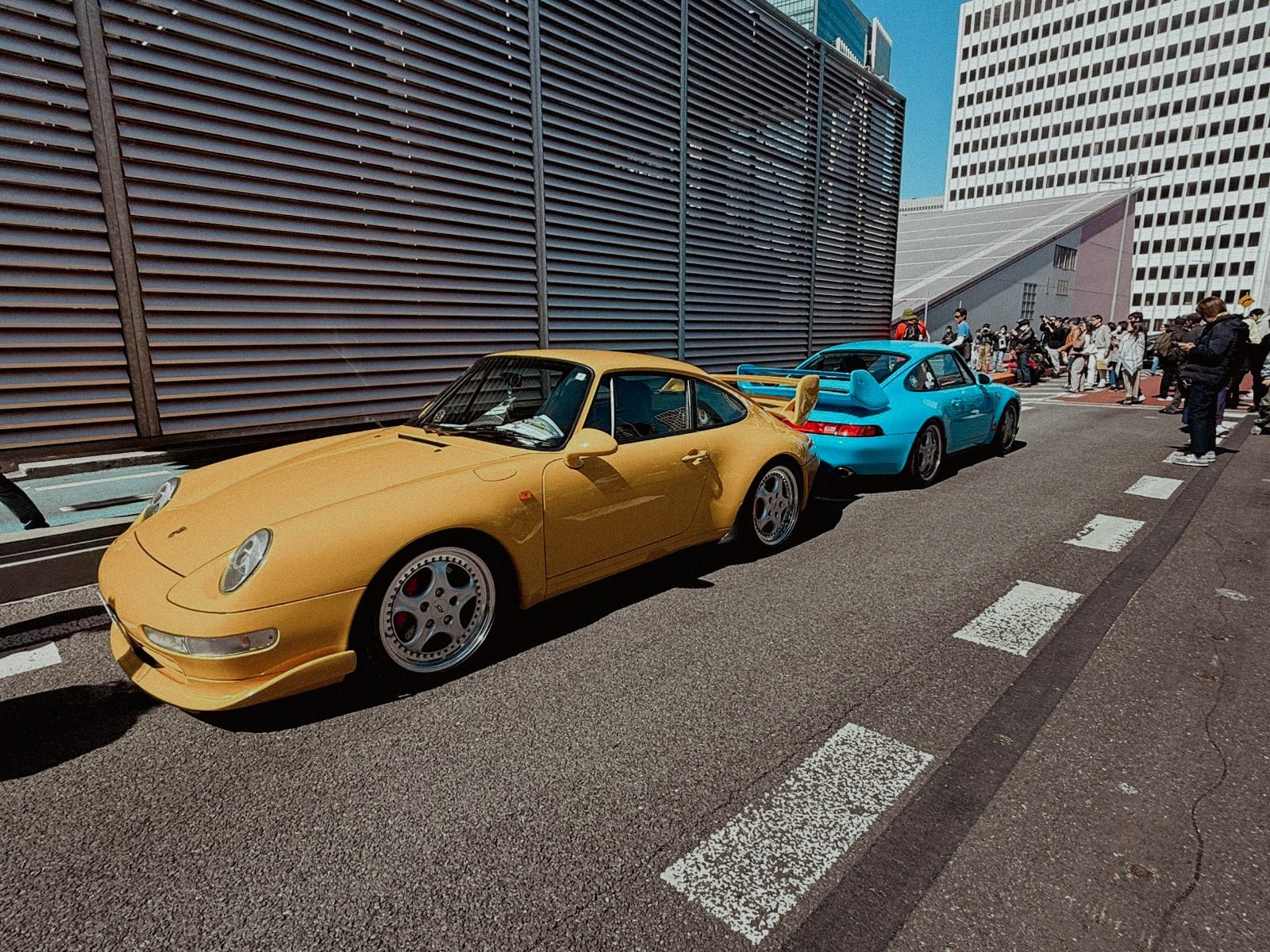 Yellow Porsche 993 GT2 and blue 993 parked side by side on the elevated expressway with crowd watching