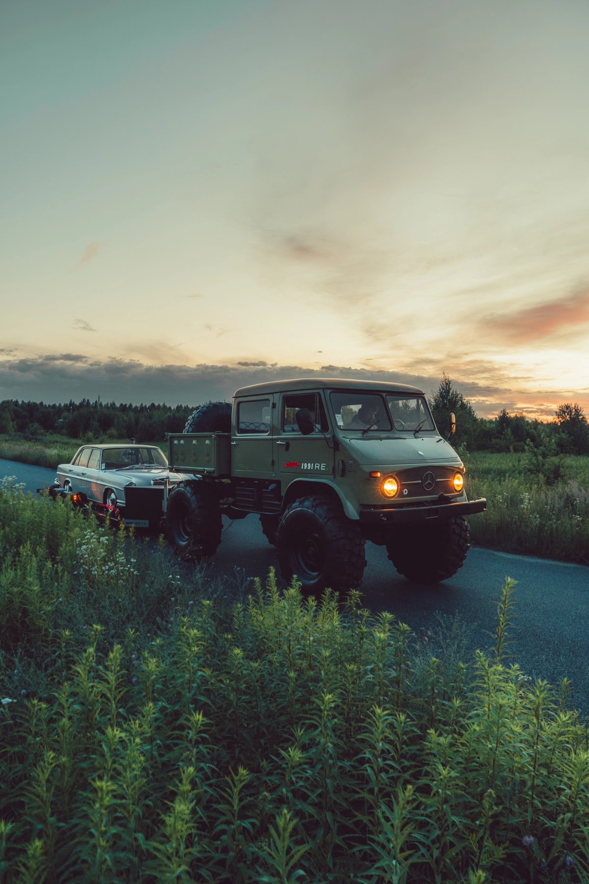Unimog with W108 on trailer on a country road at sunset with wildflowers