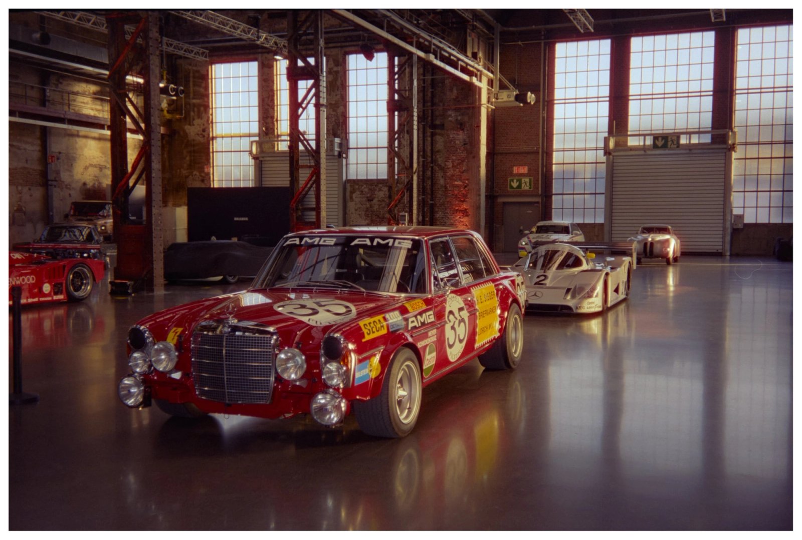 AMG 300 SEL 6.8 Rote Sau with number 35 livery in the foreground, Sauber-Mercedes C11 prototype behind it, and a red Kenwood-liveried BMW E21 race car at the far end of an Areal Böhler hall