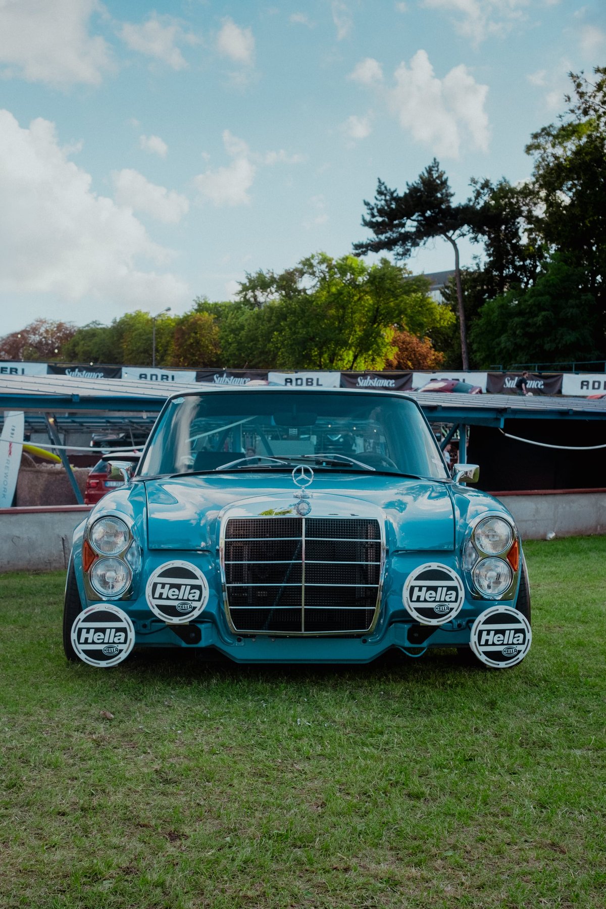 A pale blue Mercedes W114 on the grass, four Hella driving lamps mounted across its grille
