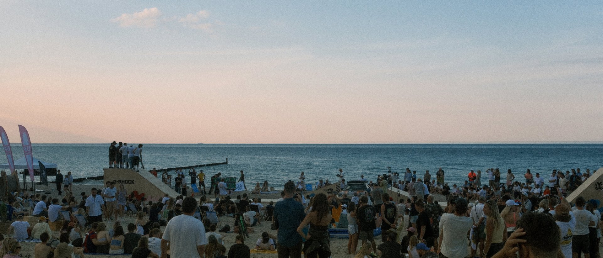 The beach at dusk during Hel Riders, a crowd on the sand around a ramp with the Baltic and a pink sky behind