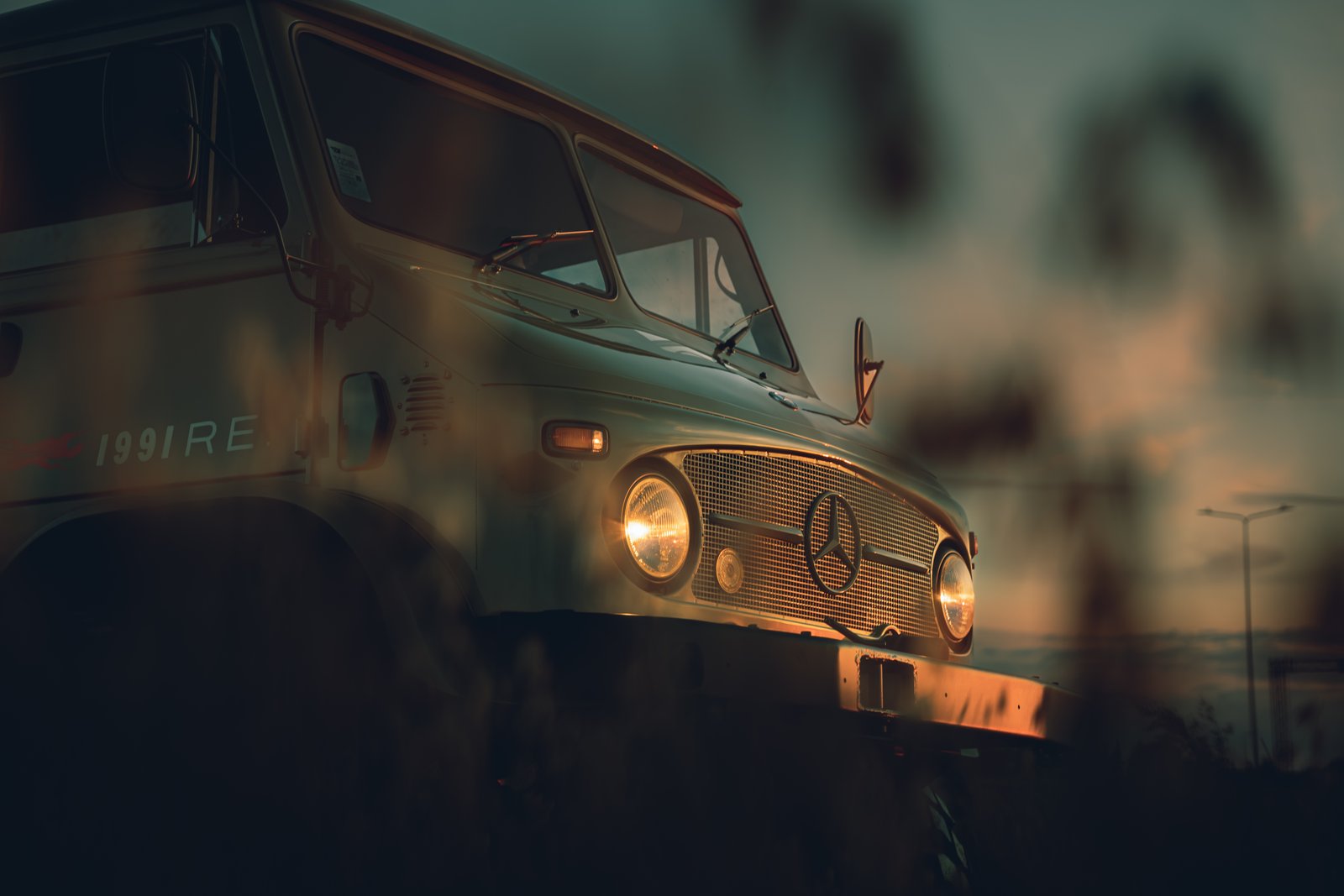 Unimog cab close-up through foliage showing Mercedes star and 1991RE lettering at golden hour
