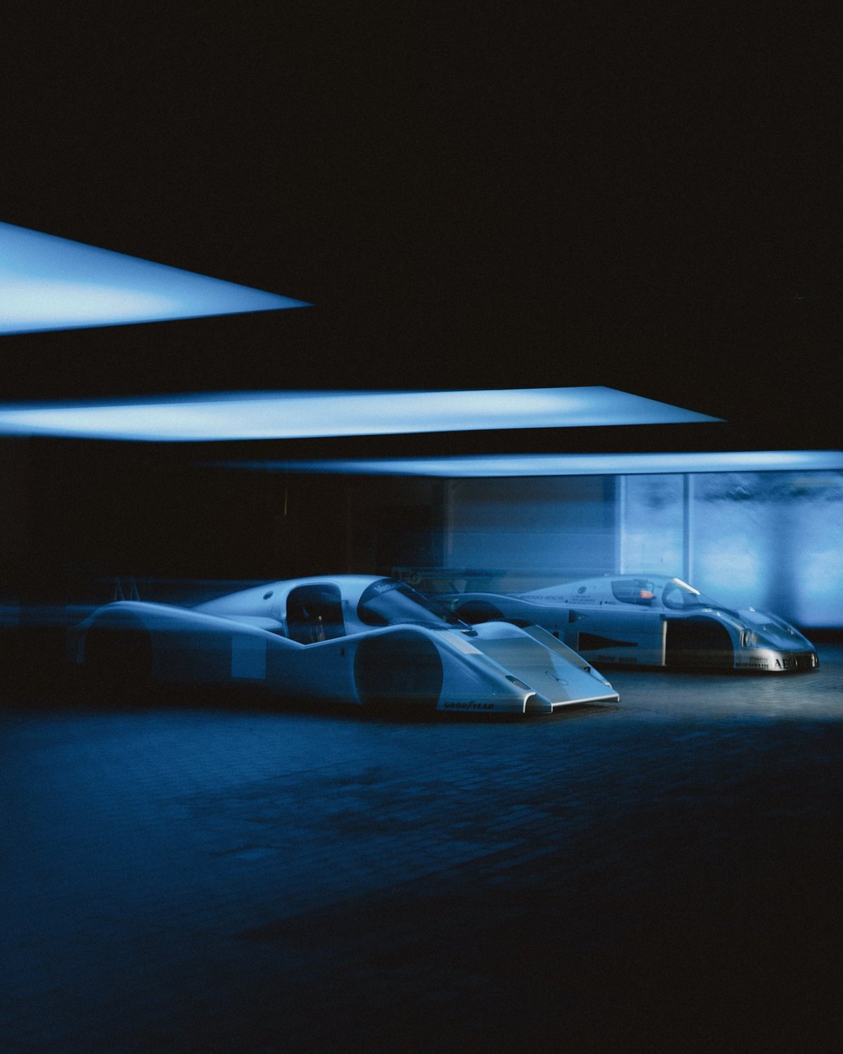 Two silver Mercedes Group C prototypes, Sauber C9 and C11, parked side by side under geometric blue-lit softboxes in a dark hall at Areal Böhler