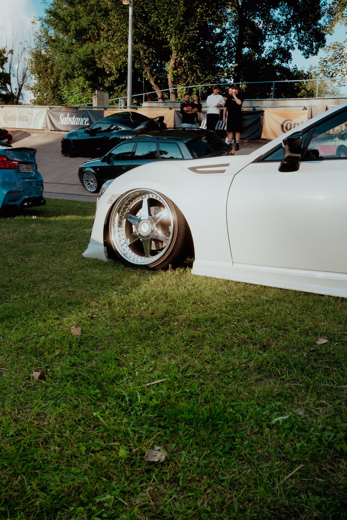 Late afternoon light on the front wheel and arch of a white widebody coupé, Substance banner on the concrete rail behind