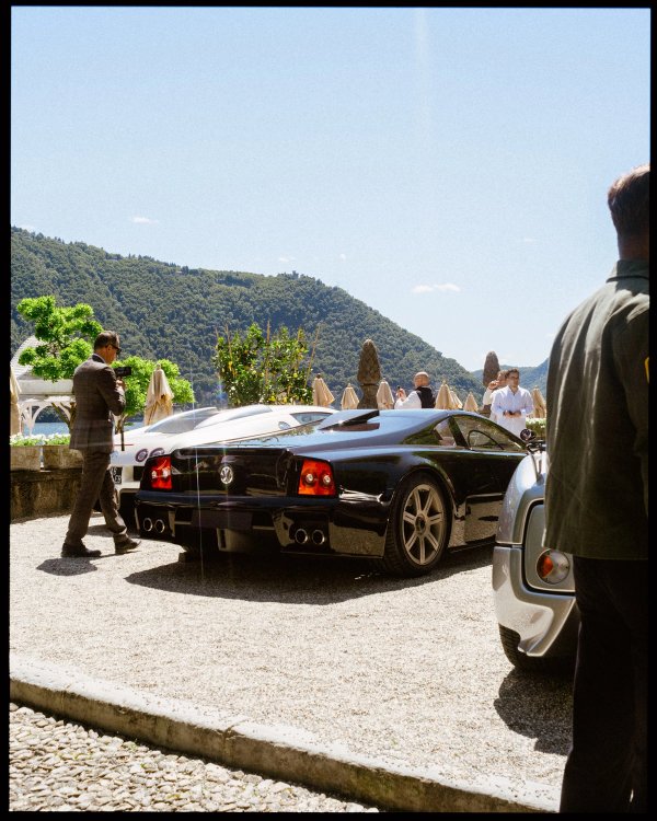 Rear of dark supercar with a man in plaid suit walking past, Lake Como hills behind