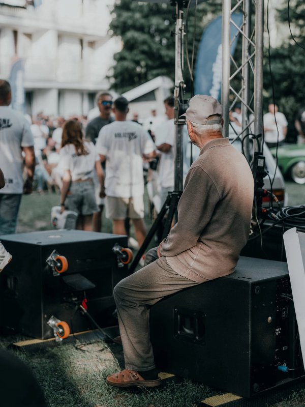 An elderly man in a cap sitting on a stage subwoofer, watching the VOLXZONE crew at work