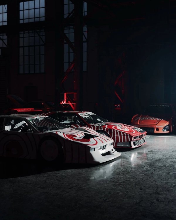 Two BMW M1 Procars with red and white spiral liveries in the foreground, orange Jägermeister Porsche Kremer K3 in the background, lit by red light in a dark industrial hall