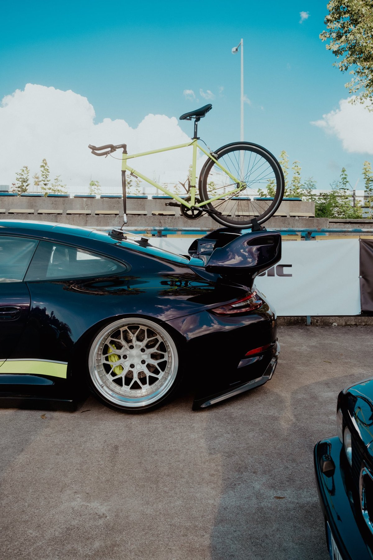 A dark Porsche 911 GT3 with a lime green fixed-gear track bike strapped to its rear wing, parked beside the velodrome