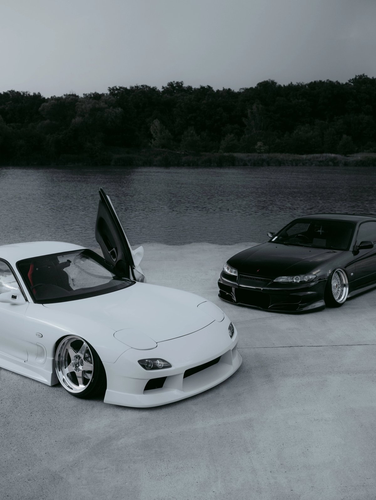 White Mazda RX-7 FD with pop-ups open and black Nissan Silvia S15 by a lake