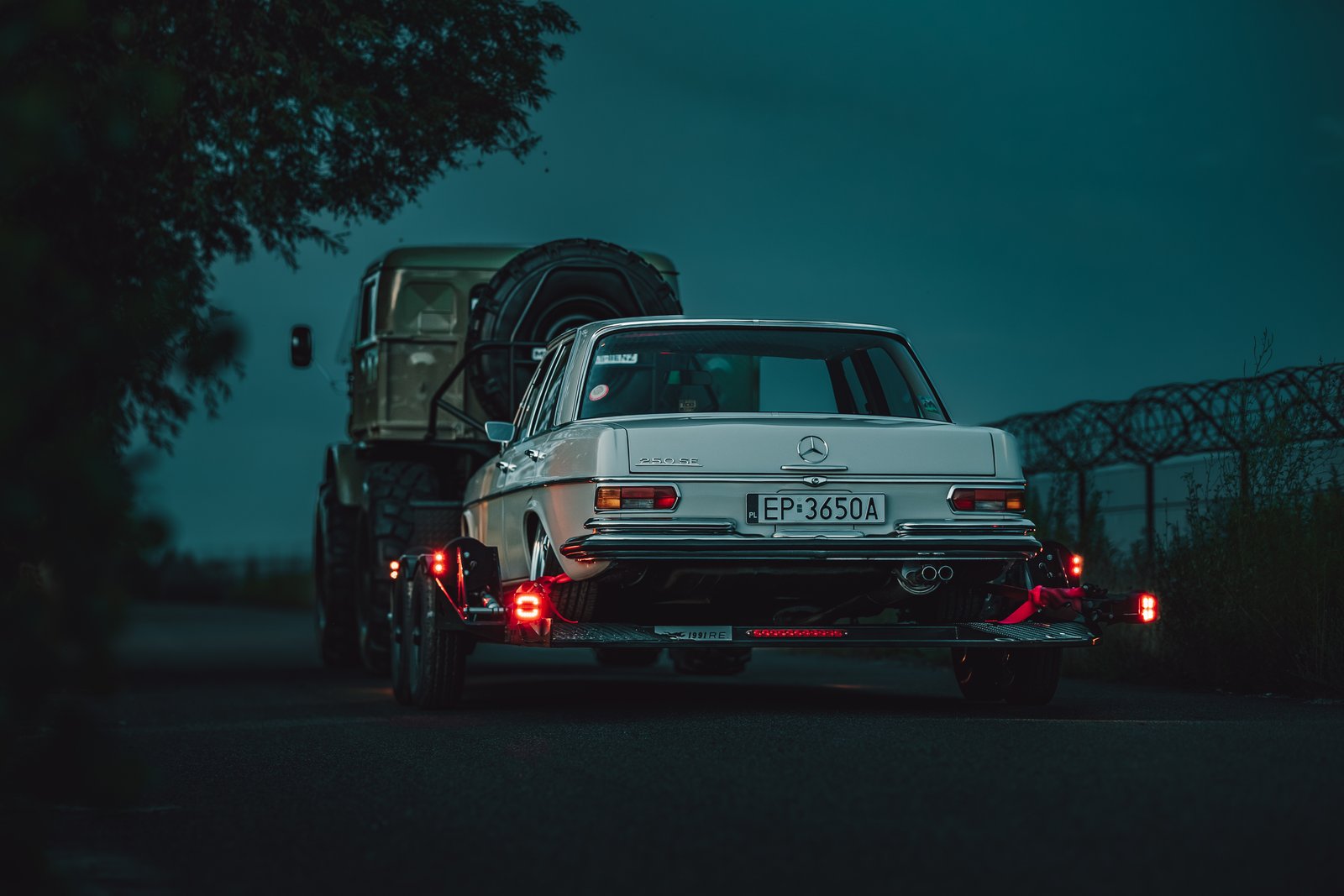 Mercedes W108 rear view on the trailer behind the Unimog at dusk with tail lights glowing