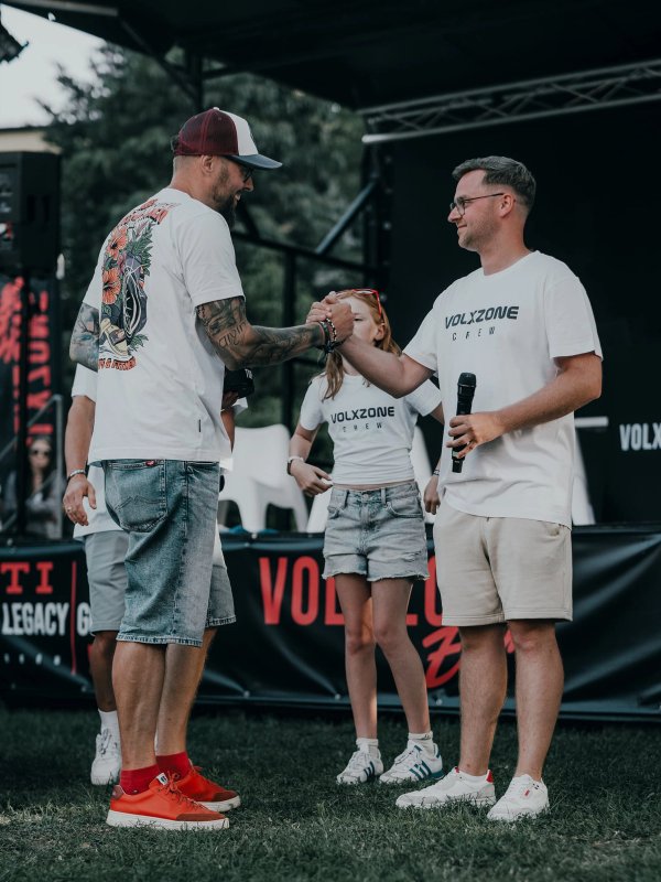 Two men clasping hands on the VolxZone stage, the crew around them in white VOLXZONE t-shirts