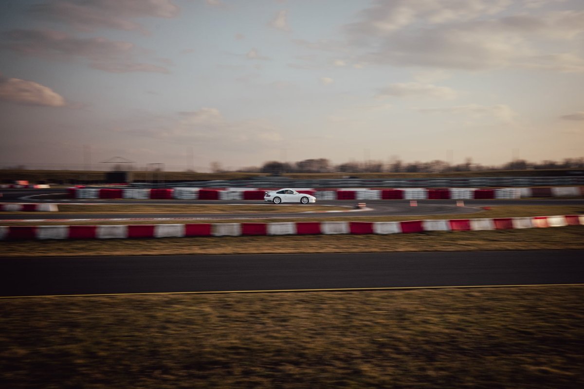 White Porsche 996.2 distant on track behind red and white barriers at dusk