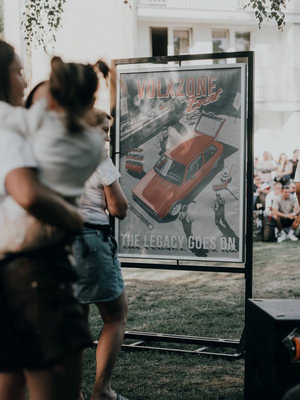 The Legacy Goes On poster on a black frame at its premiere, a mother holding a child watching it with the crowd behind