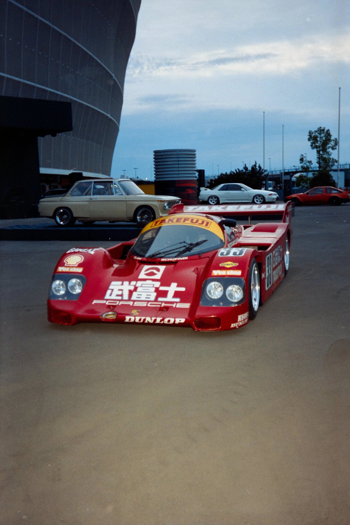Porsche 962C Team Schuppan on the esplanade at dusk with classic cars visible behind
