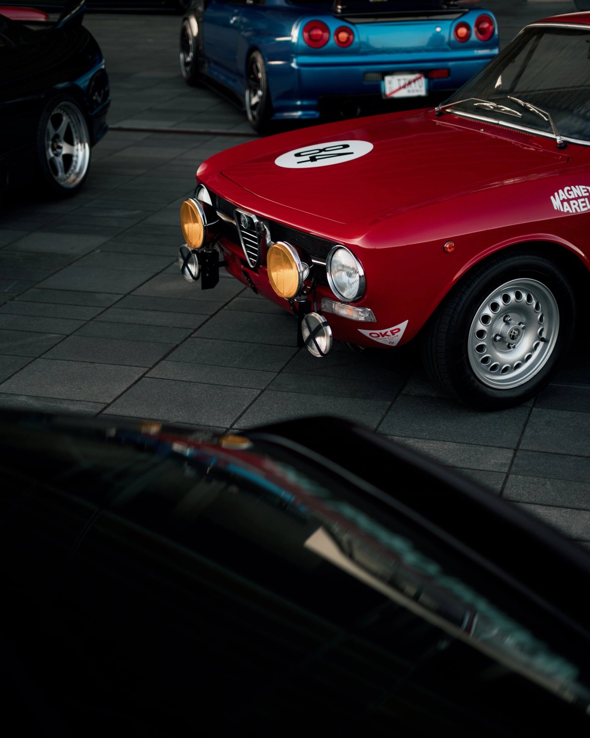 Red classic Alfa Romeo GTV with rally lights next to a blue Nissan Skyline R34 GT-R