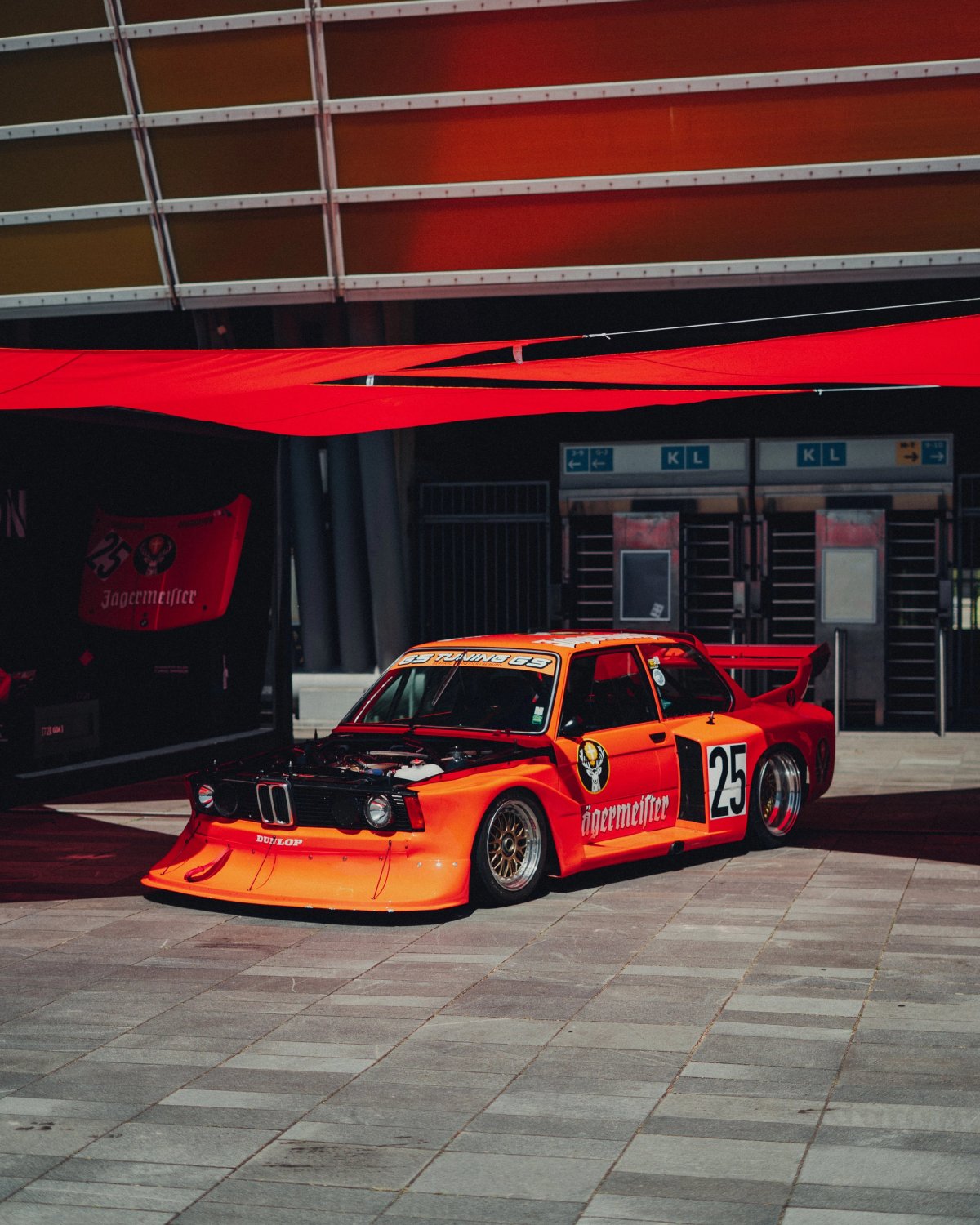 Orange BMW 320 Group 5 in Jägermeister livery, number twenty-five, under a red canopy at the venue