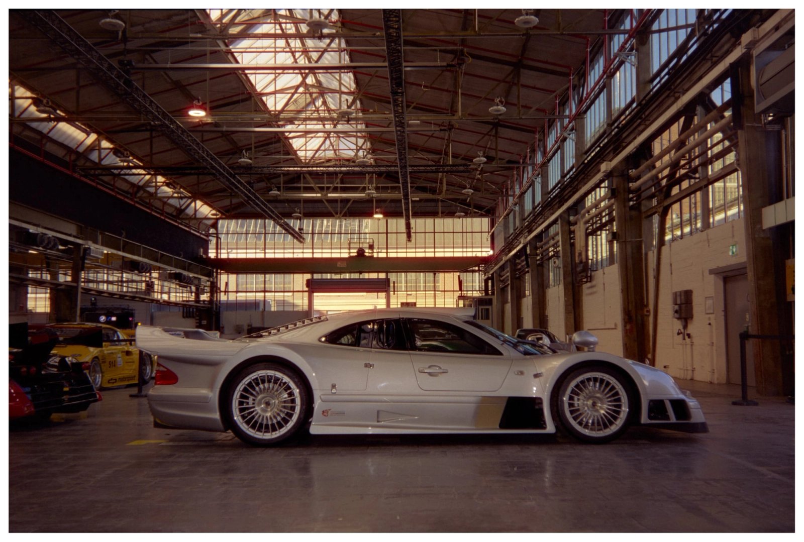Silver Mercedes CLK GTR road car in side profile inside an Areal Böhler hall, sunlight pouring through the steel-framed windows in the background