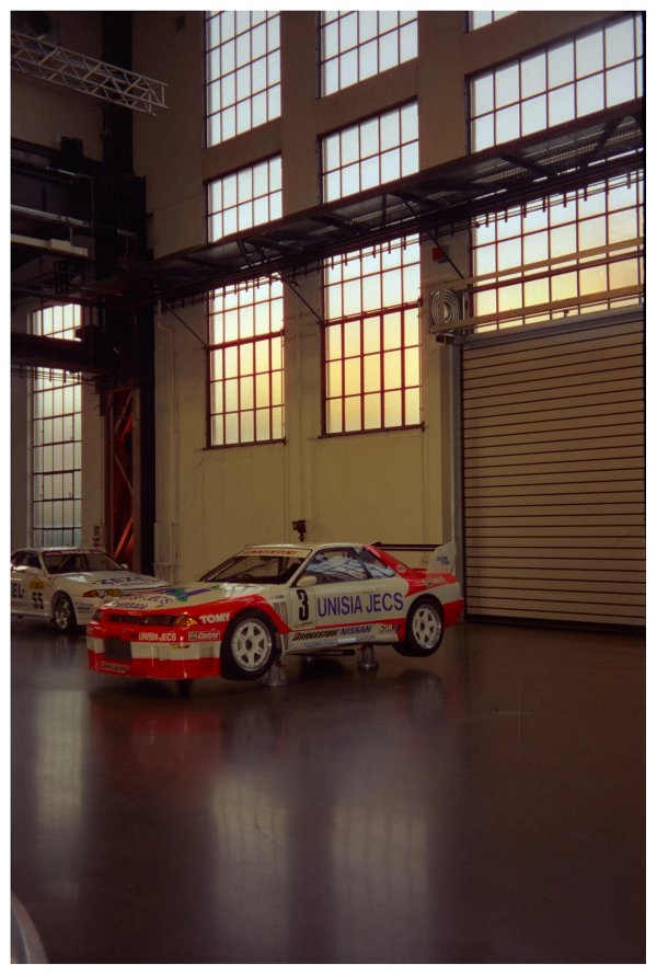 Nissan Skyline GT-R R32 Unisia Jecs in red and white livery with number 3 in the foreground, white and red Zexel R32 with number 55 behind it, inside an Areal Böhler hall with tall industrial windows