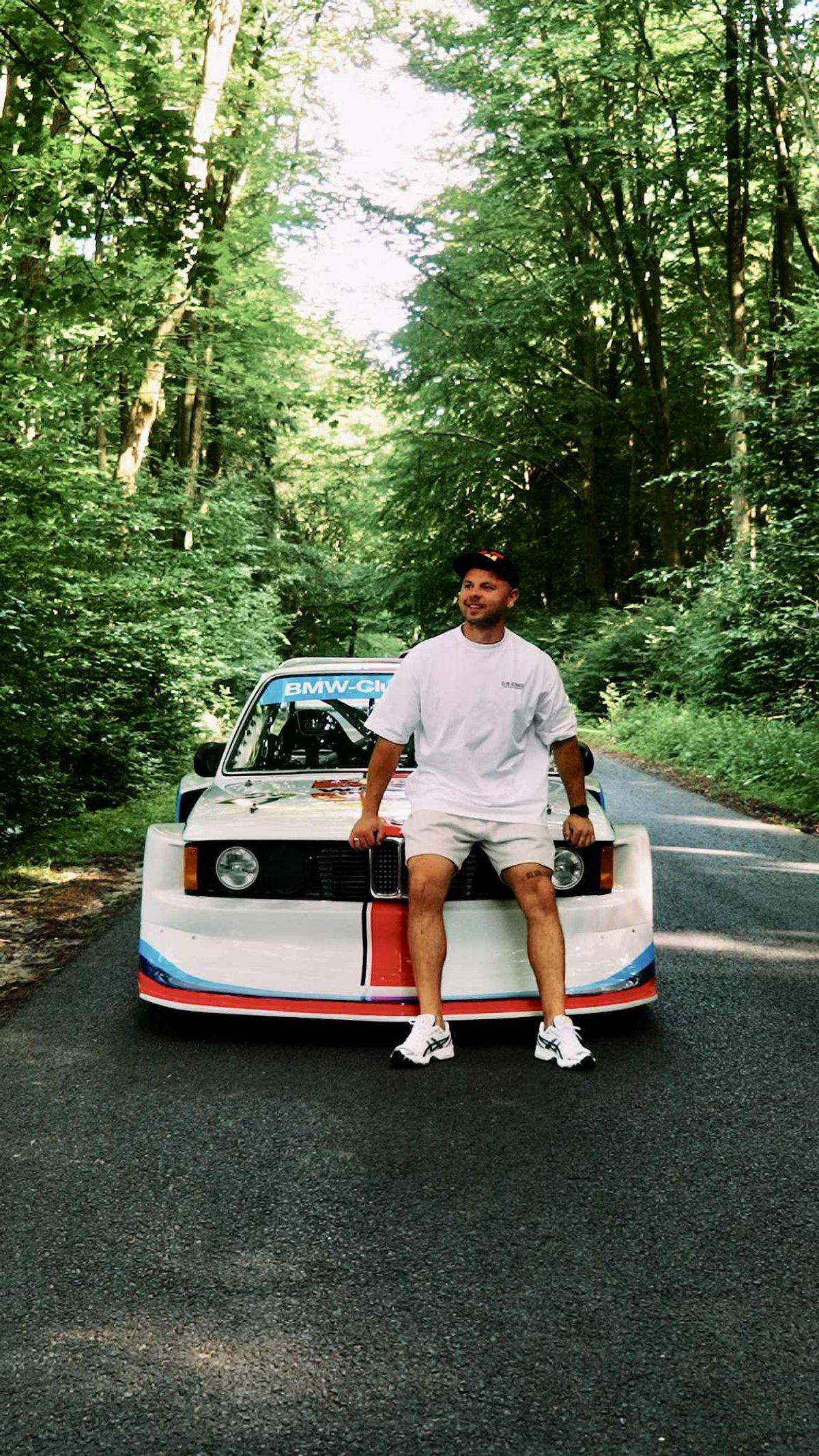 Piotrek sitting on the front of the white BMW Motorsport E21 Group 5 on a forest road