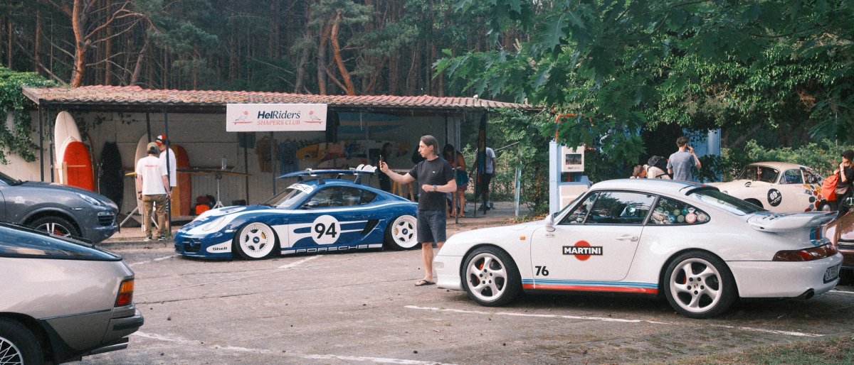 Blue and white Porsche 964 number 94 and a white 993 in Martini livery number 76 by the HelRiders banner, a man photographing them
