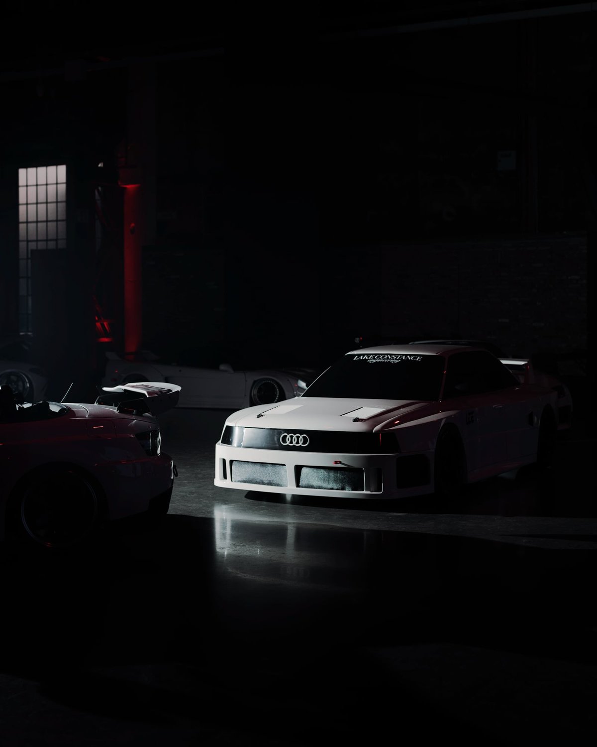 White Audi 80 quattro with Lake Constance Engineering windshield banner and a Honda S2000 in the foreground inside a dark industrial hall lit by faint red column light