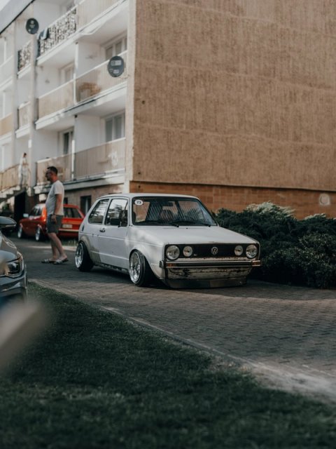 White patinated Golf Mk1 with race number 73 parked between the apartment blocks, a man in slides walking past