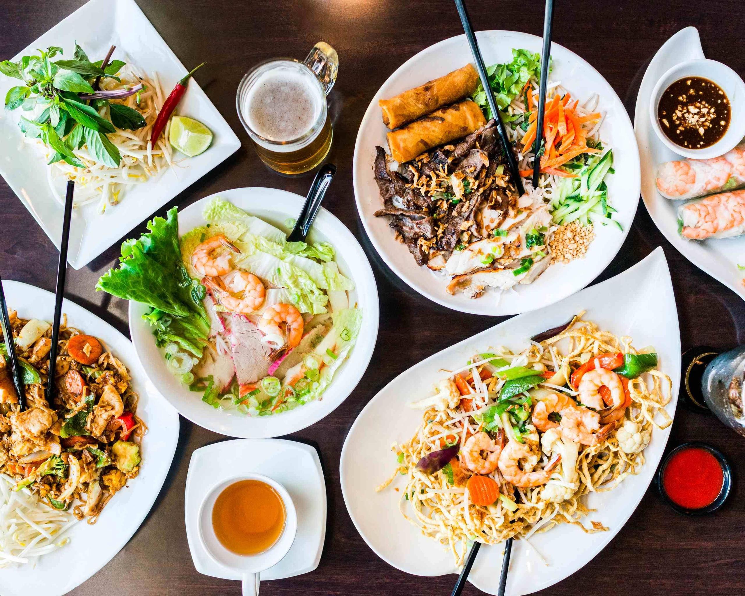 A top-down overhead view of a Vietnamese feast featuring shrimp noodle soup, stir-fried noodles, spring rolls, and grilled pork vermicelli on a dark wood table.