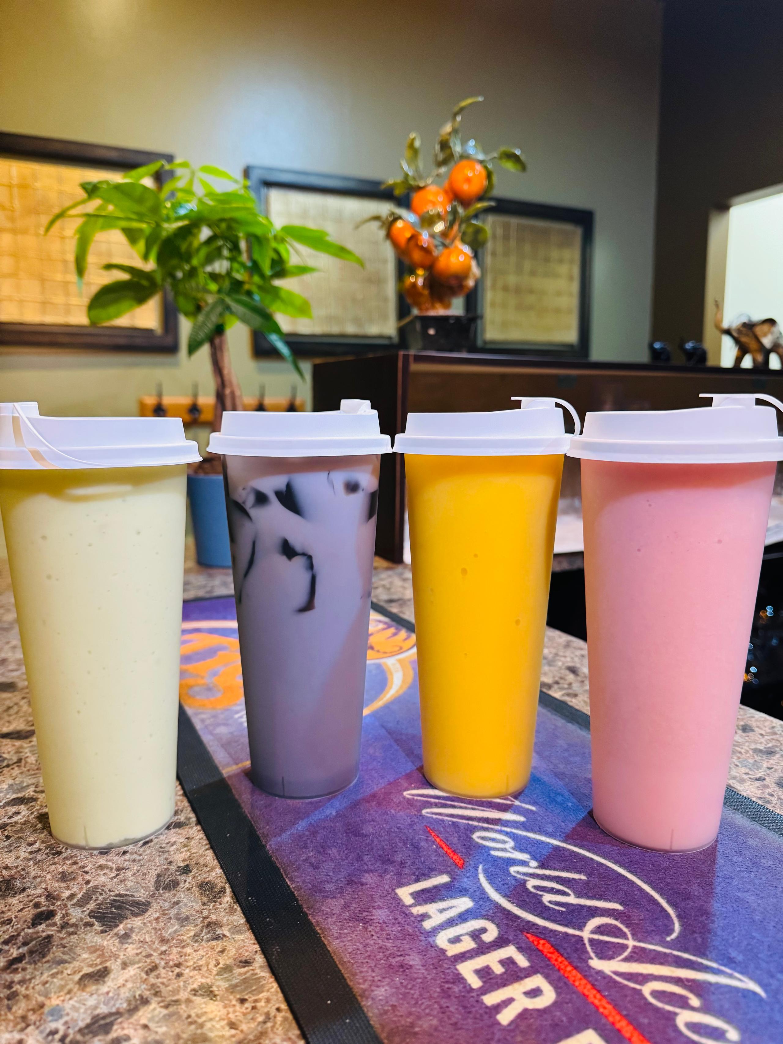 A row of four colorful fruit smoothies and bubble tea drinks in tall cups with white lids, including avocado, taro with grass jelly, mango, and strawberry flavors.