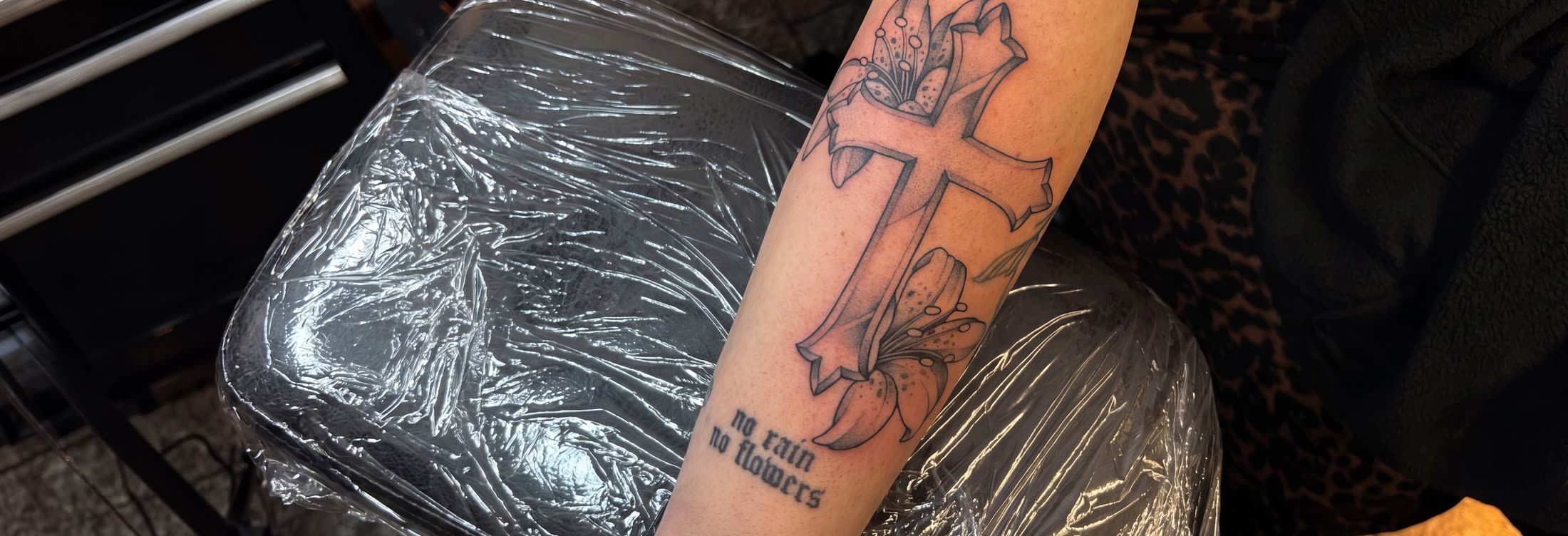 Kreuz mit Lilie und Schriftzug ‚no rain no flowers‘ am Unterarm — Fineline-Tattoo von Melinda Dür, Vorarlberg