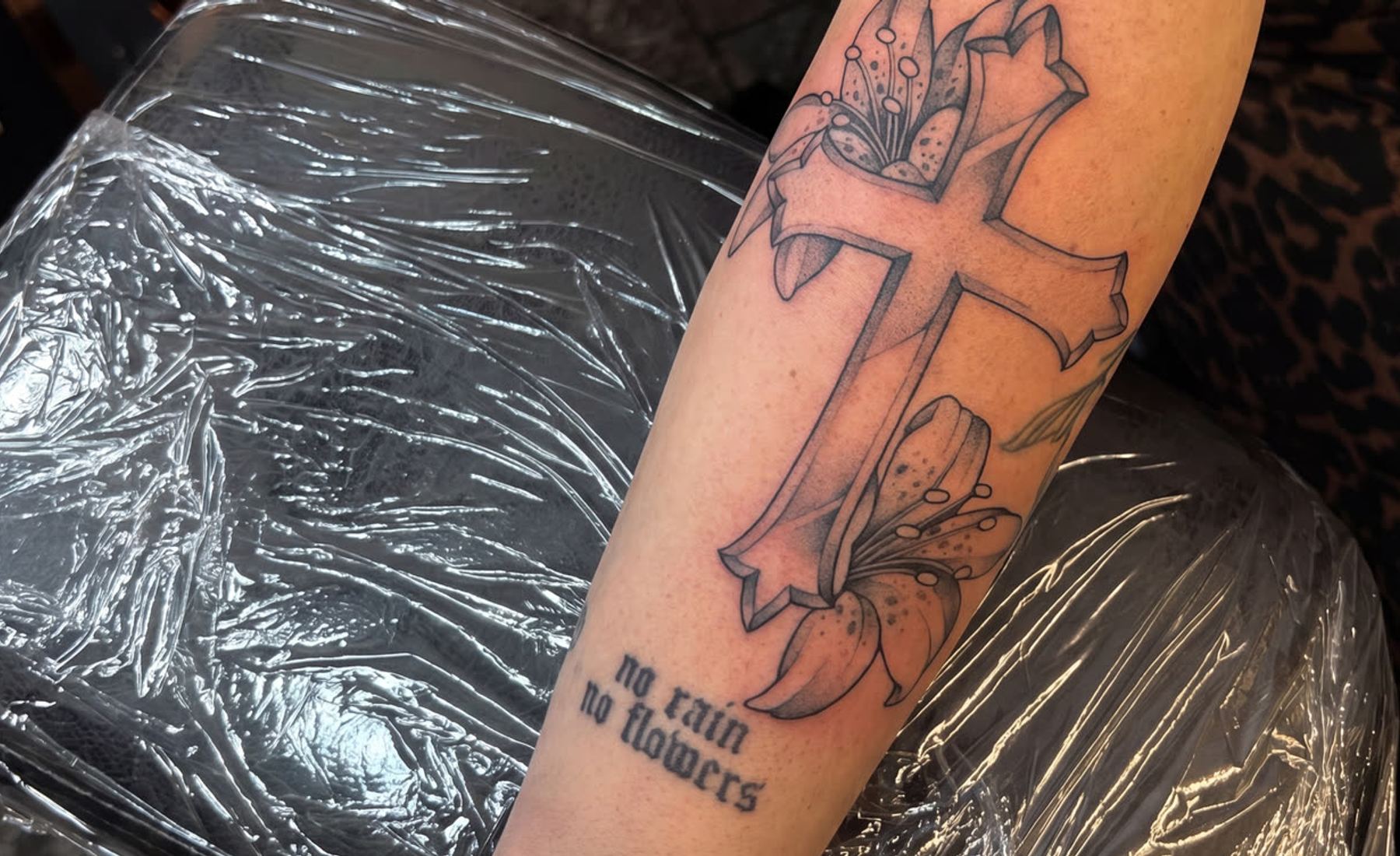 Kreuz mit Lilie und Schriftzug ‚no rain no flowers‘ am Unterarm — Fineline-Tattoo von Melinda Dür, Vorarlberg