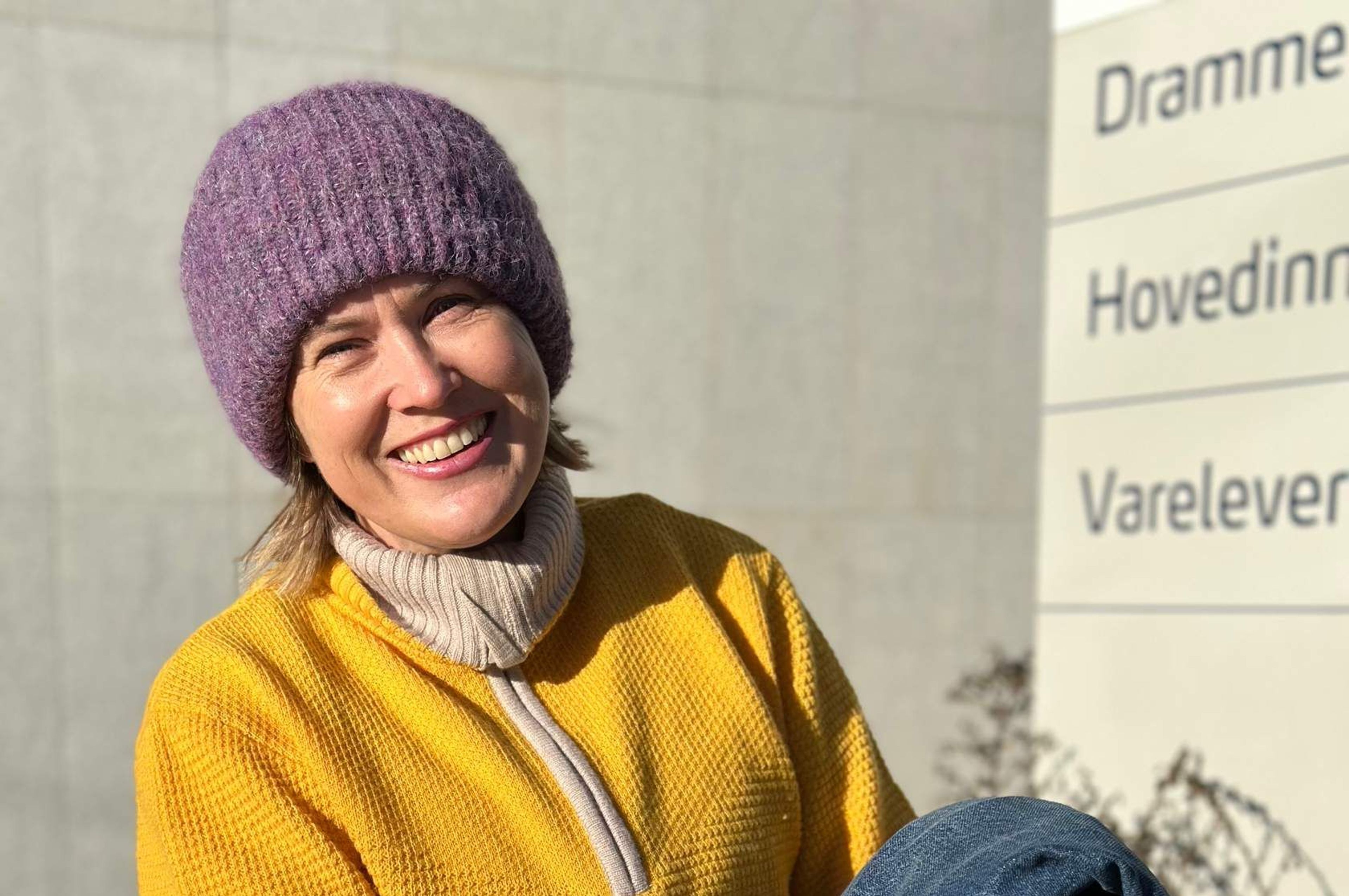 Kvinne i gul genser og lilla lue som smiler mot kamera