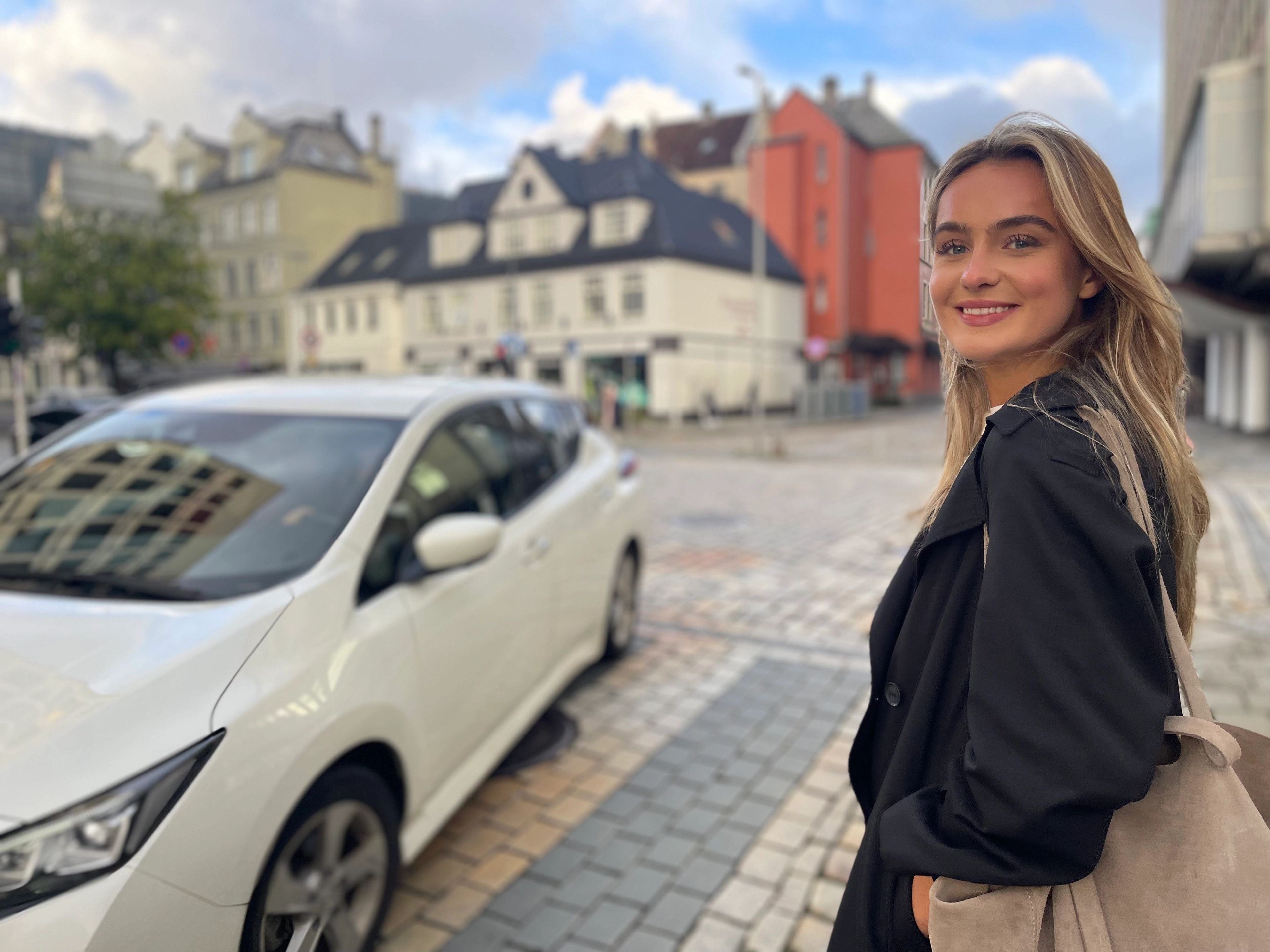 Person som ser smilende inn i kamera med en bil som krysser veien foran henne