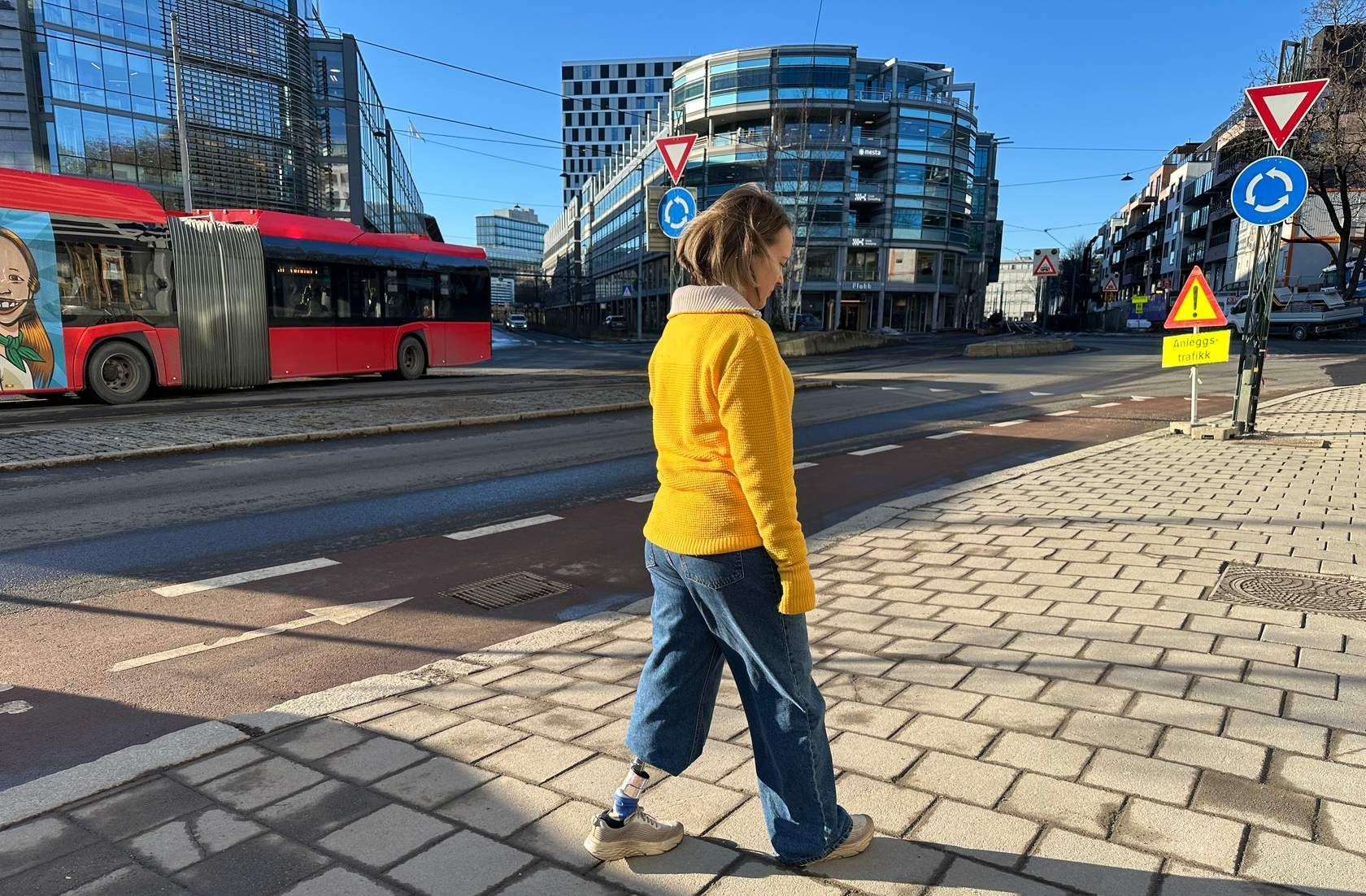Person med benprotese som går på fortauet med en buss ved siden av