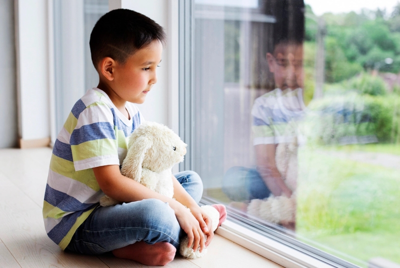 Liten gutt sitter og ser ut av vinduet, med kosebamse i armene.