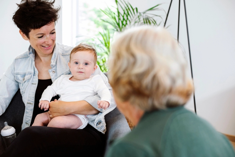Baby sitter på fanget til mor. Familiekontakt sitter med ryggen til.