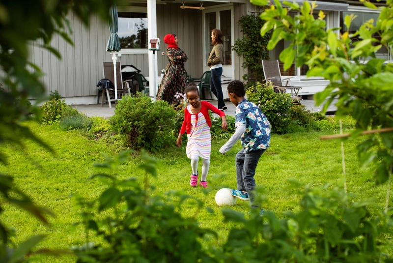 Barn spiller fotball. Mor og familiekontakt i i samtale i bakgrunnen.