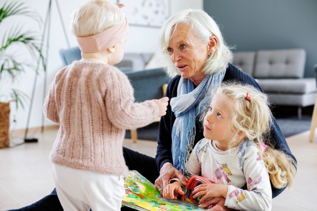Liten jente står med med ryggen til. Familiekontakt og storesøster sitter på gulvet med bok.