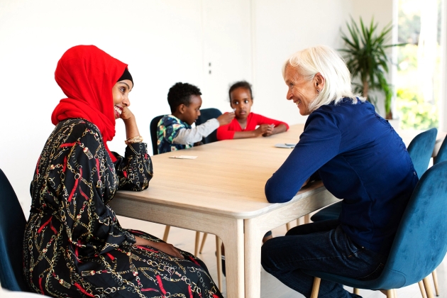 Familiekontakt og mor sitter ved bord og smiler til hverandre. To barn sitter sammen ved dem ved bordet.