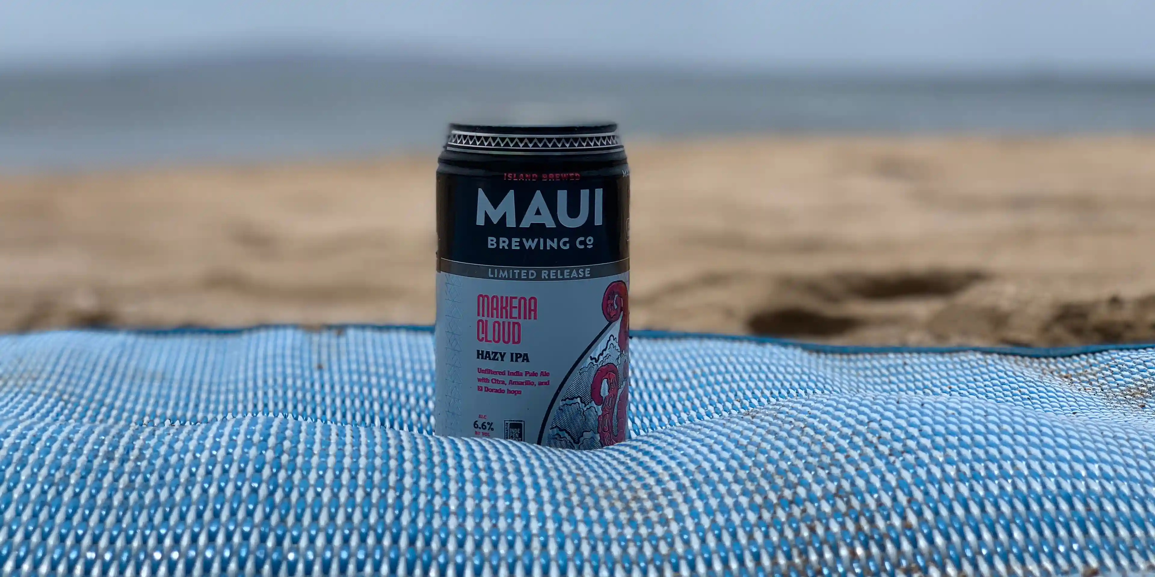 Shot of a can of Maui Brewing Co beer, on a blanket, with the beach in the background.