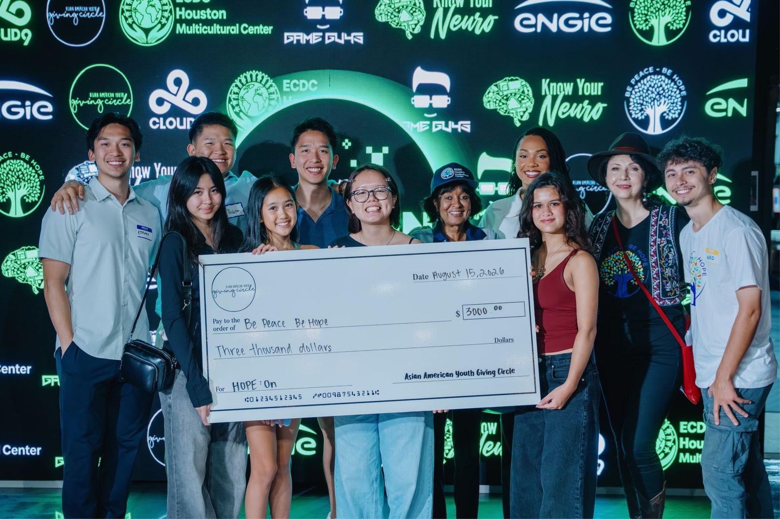 Young people, partners and the Be Peace – Be Hope team holding the Asian American Youth Giving Circle’s check for HOPE:ON in front of the sponsors’ wall at Game Guys