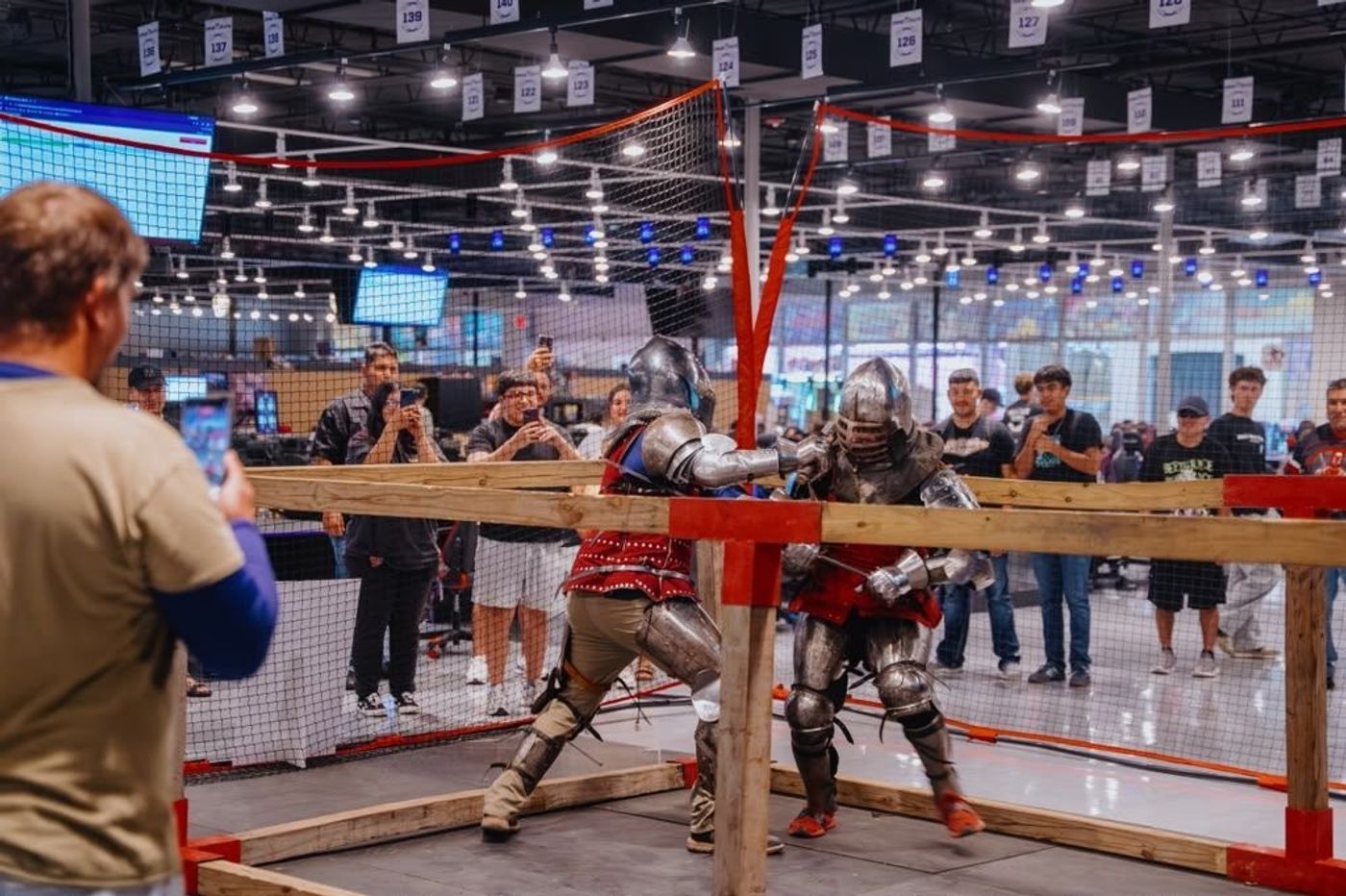 Two fighters in full armor face off in the arena as guests film
