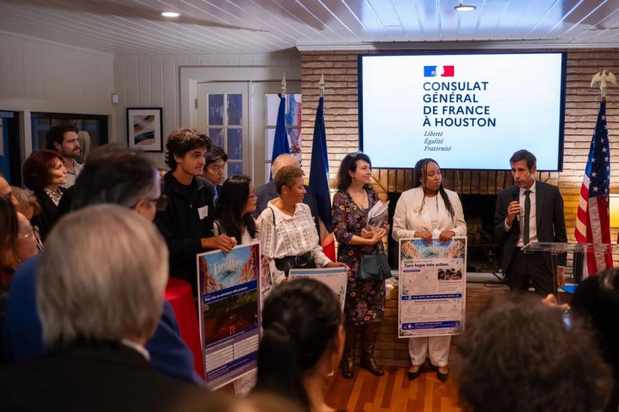 A speaker addresses guests while young people hold EsperHope posters beside the Consulat Général de France à Houston screen