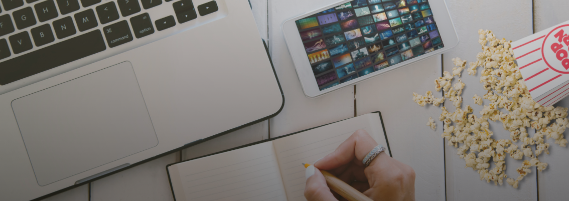 Top-down view of a workspace with a laptop, popcorn, and a person writing notes from Netflix, symbolizing a productive way to learn business from movies.