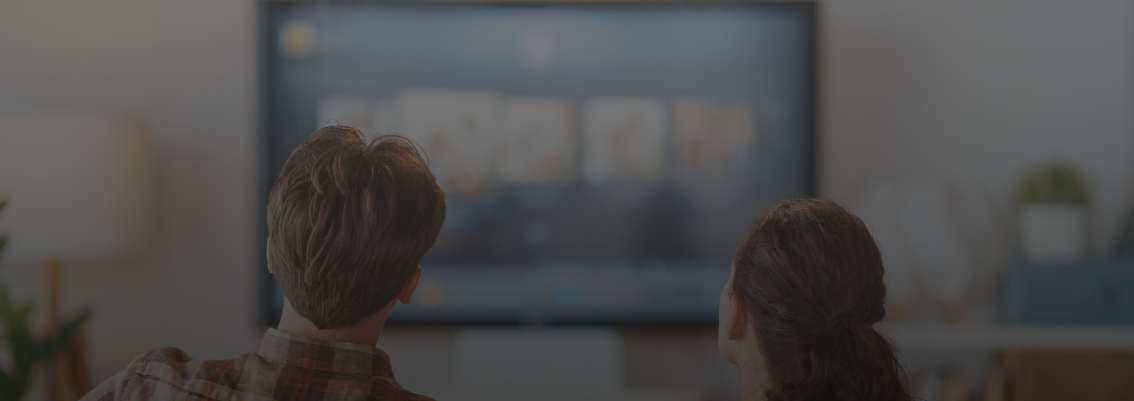 Two people, a man in a plaid shirt and a woman with dark curly hair, sitting on a sofa from behind, looking up at a blurred large screen TV in a living room, suggesting they are watching a movie or TV show together.