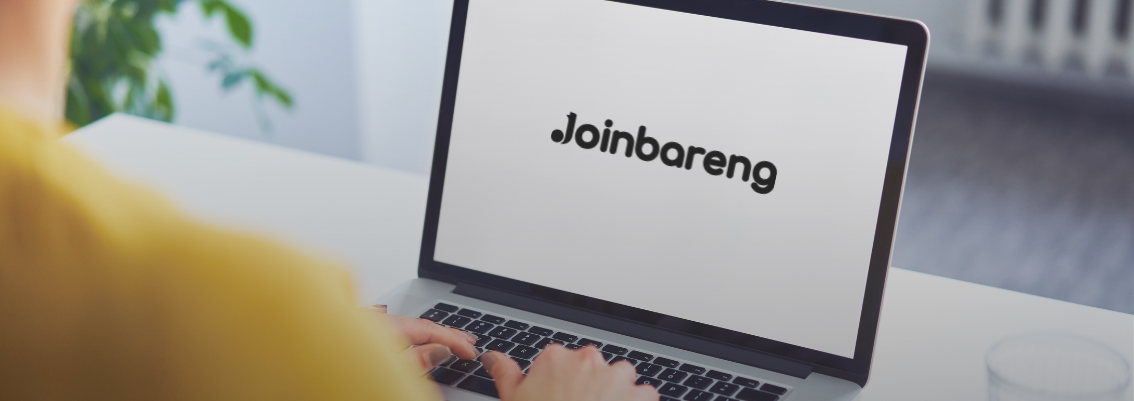 A person from behind, wearing a yellow shirt, typing on a silver laptop with the "Joinbareng" logo displayed prominently on the screen. A glass of water is on the white desk next to the laptop, and a blurred bright room with a plant is in the background.