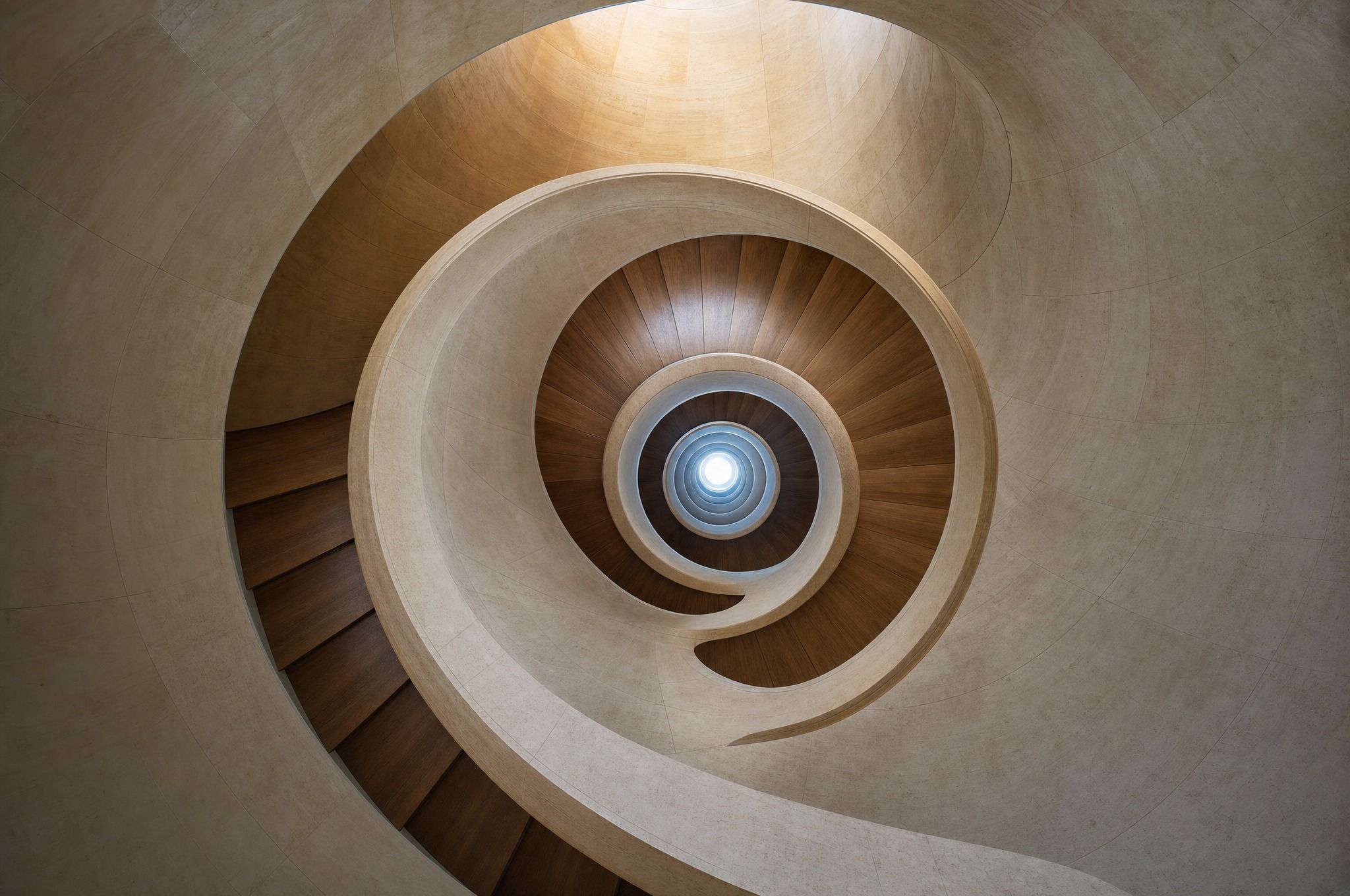 Looking up the centre of a curving staircase with timber treads and smooth pale stone walls, its spiral winding towards a small round skylight.