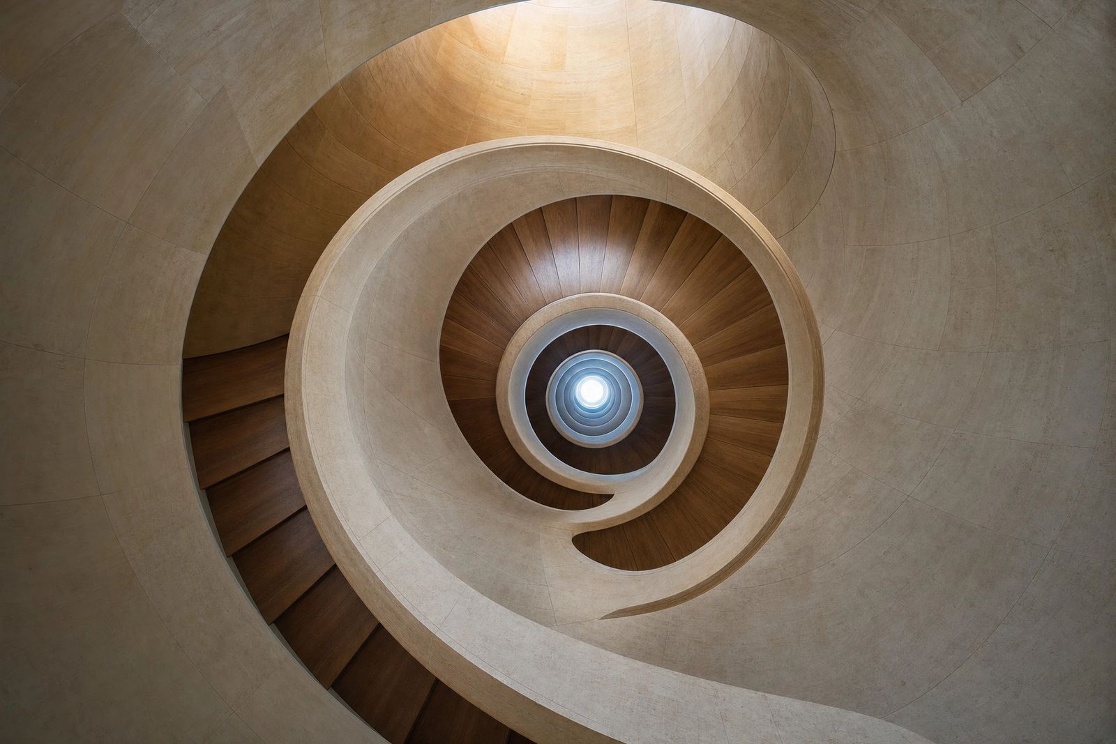 Looking up the centre of a curving staircase with timber treads and smooth pale stone walls, its spiral winding towards a small round skylight.