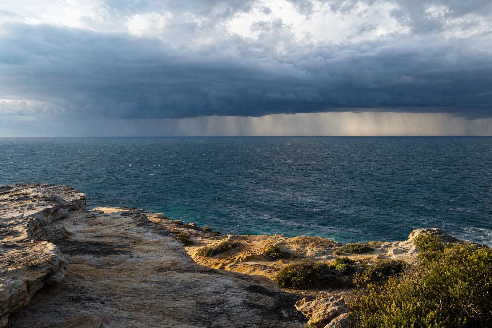 A dark band of storm cloud and rain gathers over the Pacific Ocean on the horizon, seen from a sunlit sandstone headland with coastal scrub in the foreground.