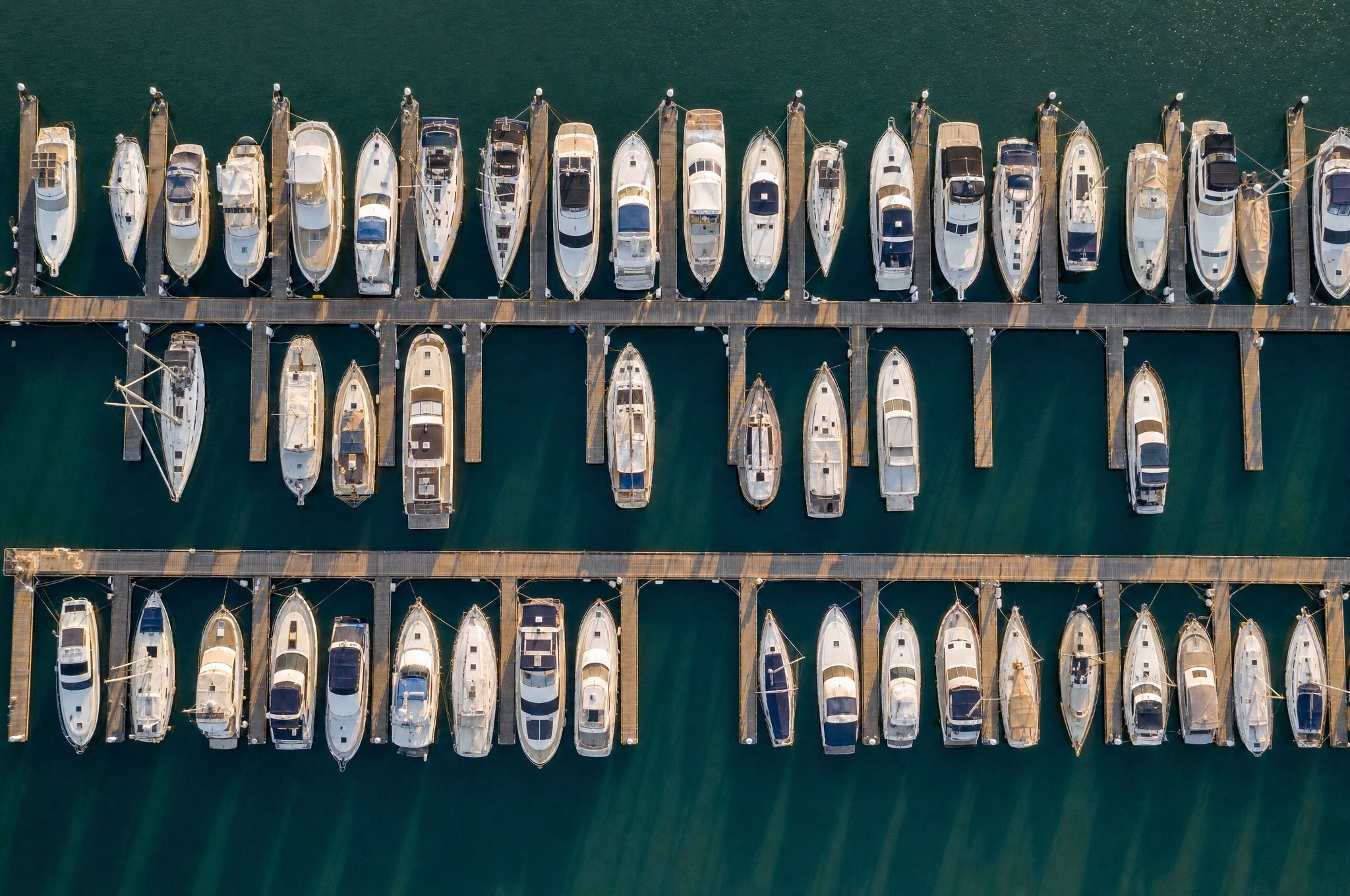 An aerial view looking straight down on a calm marina, with rows of moored yachts and motor cruisers in their berths along timber finger jetties on deep green water.
